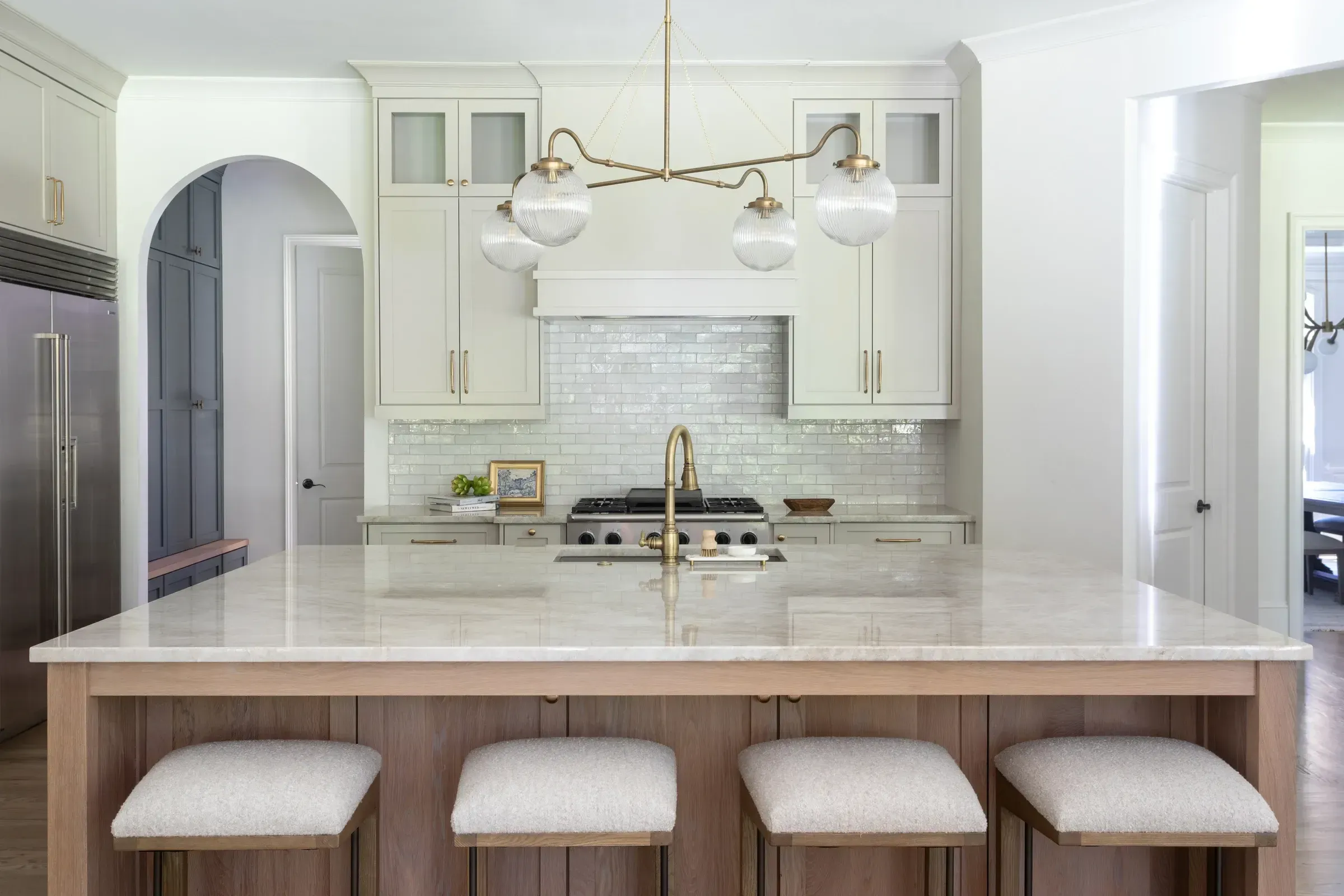 Bright white kitchen with natural wood accents, brass fixtures, a textured backsplash, and a spacious island.